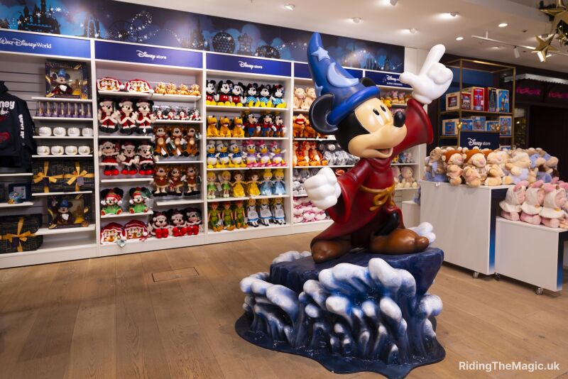 statue of Mickey mouse in sorcerer outfit with rows of soft toys behind blue signs with white writing above soft toys laminate floor