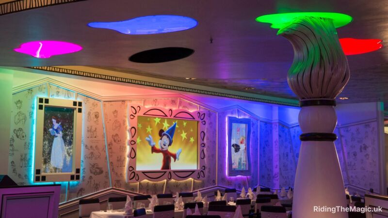 large white paint brush floor to ceiling colour splotches lights on ceiling, black and white seating and white table bottom of picture walls have 3 painting from left to right: snow white, mickey mouse, cartoon ostrich with pink bow on head.