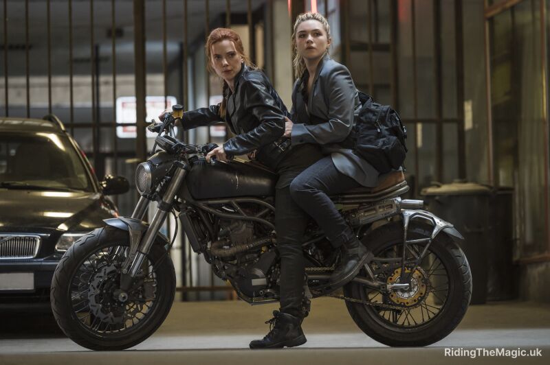 woman with red hair (black widow) in black clothes on black motorbike with woman with blonde hair sitting behind in black clothes