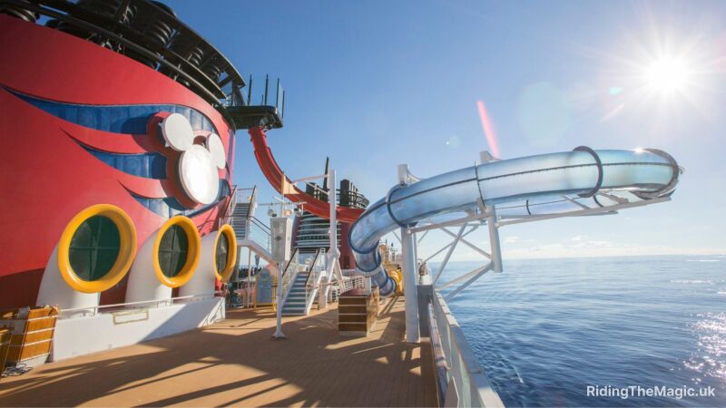 clear tube elevated above wooden cruise ship deck reachingo ut over blue sea with blue sky backdrop red funnel to right and small flight of white stairs. Funnel has white mickey mouse face silhouette and 3 yellow rimmed port holes