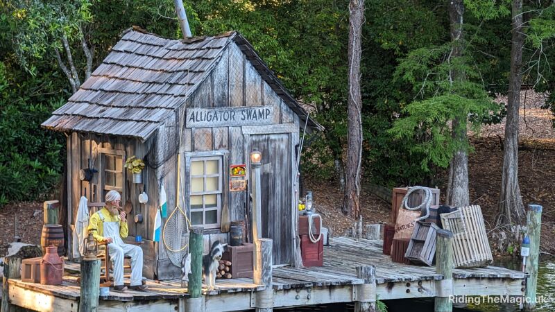 A wooden shack by the river with a old man sitting outside with his dog. The shack has a sign which says Alligator Swamp