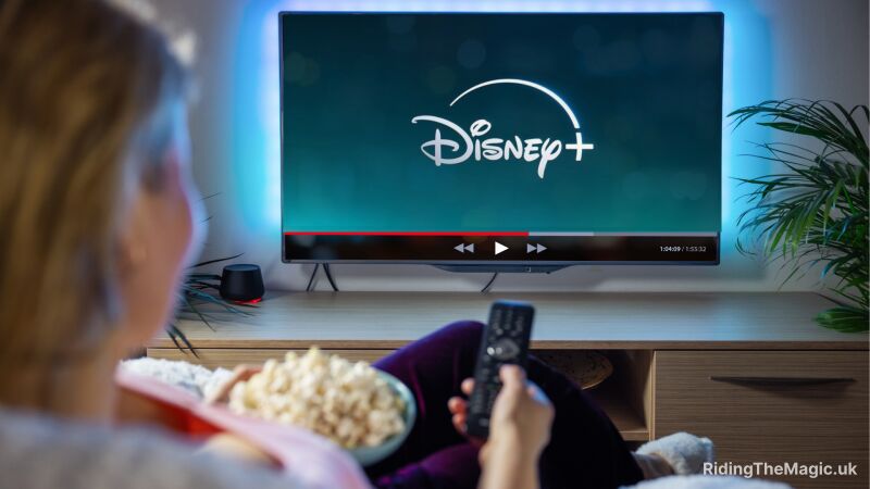 back of blonde womans head blurred pointing black remote at TV with Disney+ logo on screen. TV in centre on unit