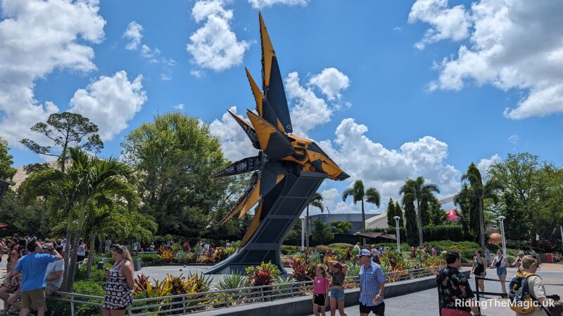 Grey and yellow Nova Corps Starblaster spaceship surrounded by flowers and a silver fence with bench blue sky people walking round