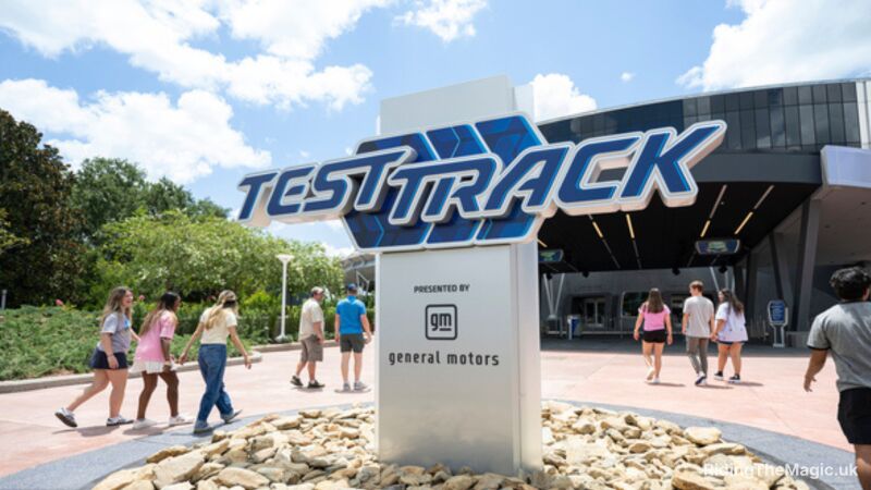 grey sign rising from circular roundabout in pavement full of beige stones, Blue sign with words Test Track people walking either side ride building behind
