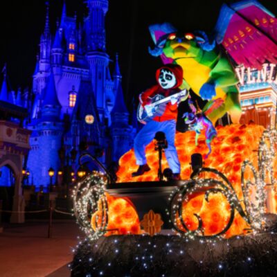 Nighttime parade float with a colorful dragon and a skeleton musician glowing in orange and blue lights in front of a lit castle.