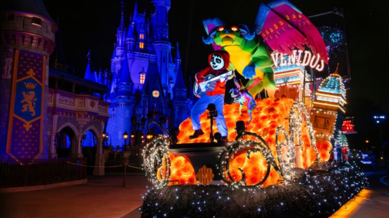 Nighttime parade float with a colorful dragon and a skeleton musician glowing in orange and blue lights in front of a lit castle.