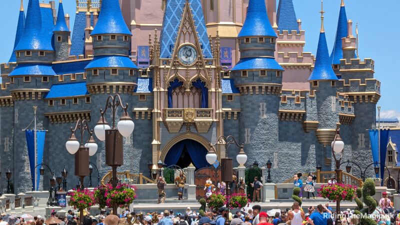 grey castle with blue tiled turrets and pink building behind lamp posts with two orb white lights hanging baskets pink flowers tops of peoples heads in crowd