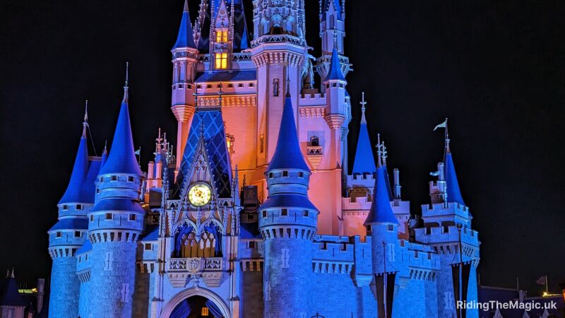 A brightly lit fairytale castle illuminated in blue and pink against a night sky.