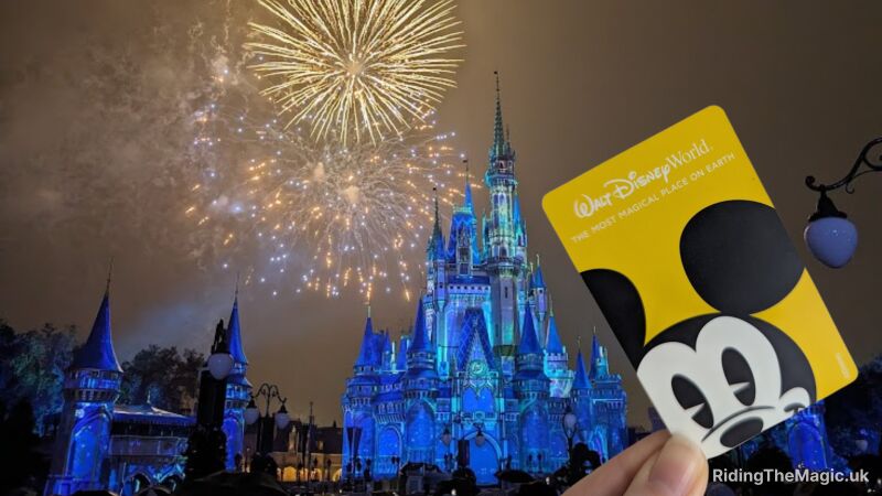 Fireworks explode over the blue‑lit Cinderella Castle at Walt Disney World as a hand holds a Mickey-themed park card in the foreground.